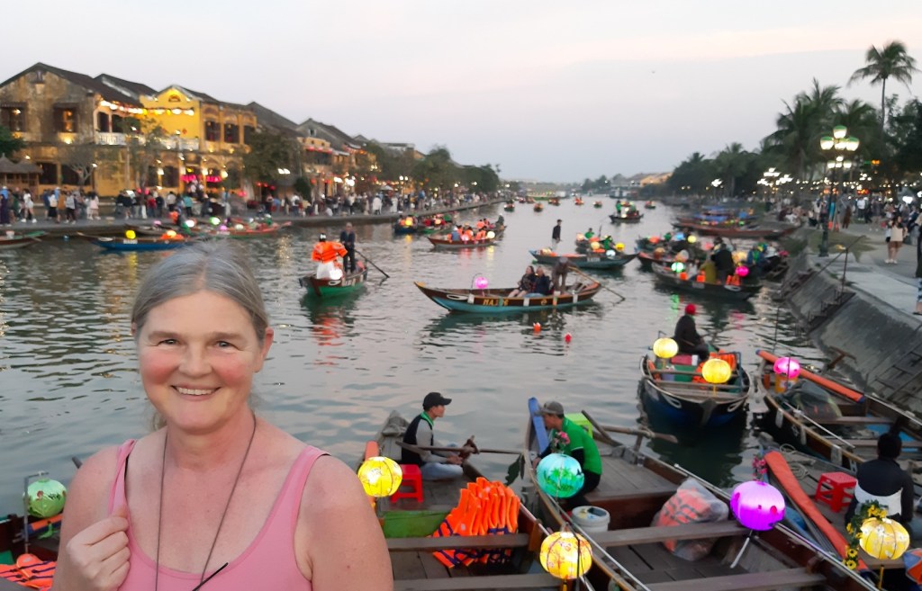 Die Ergotherapeutin Katja Hausmann lächelt glücklich in die Kamera. Sie steht in Hoi An, einer Stadt in Vietnam, auf einer Brücke im Abendlicht. Im Hintergrund schwimmen viele Boote auf dem Fluss. Sie sind mit den bunten Lampignons, die die Stadt Hoi An berühmt gemacht haben, reich geschmückt.